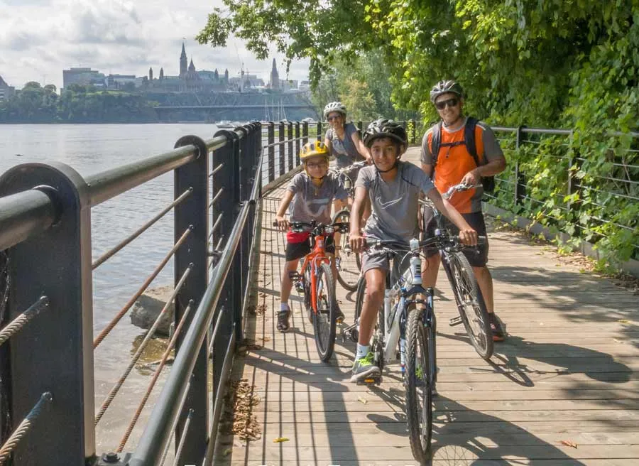 Parents and kids cycling on a wooden bridge with the view of Ottawa river and parliament building on an Ottawa express tour